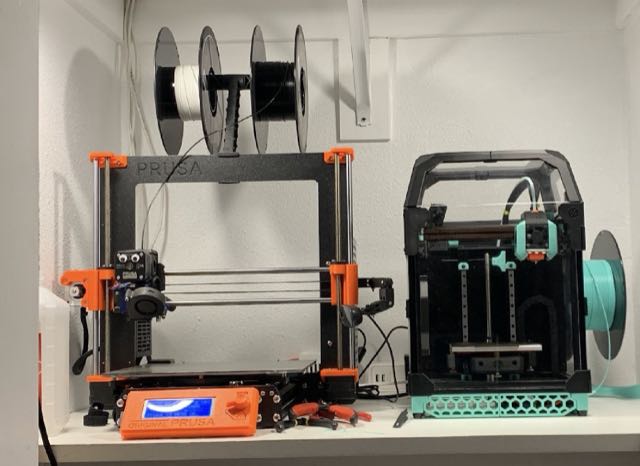 Prusa Mk3S (left) and Voron 0 (right) 3D printers side by side on a shelf, with filament spools mounted above