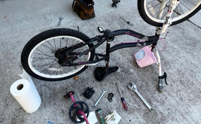 Kid’s bike partially disassembled on a driveway for repair, with crank, pedals, and tools laid out