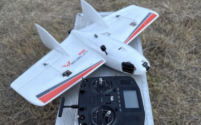 White foam flying-wing UAV (Nano Goblin) resting on its RadioMaster transmitter, on dry grass
