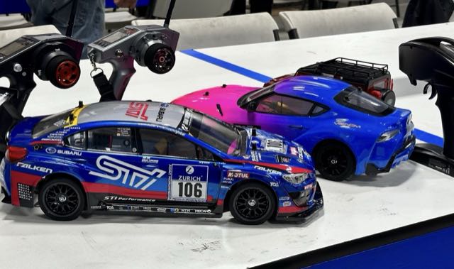 Two TT-02 RC touring cars with Subaru WRX STI and Toyota Supra bodywork, sitting on a table next to their transmitters