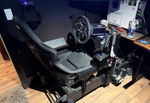 Full sim-racing rig with aluminum profile frame, racing seat, direct-drive wheel, shifter, handbrake, and a VR headset hanging beside it